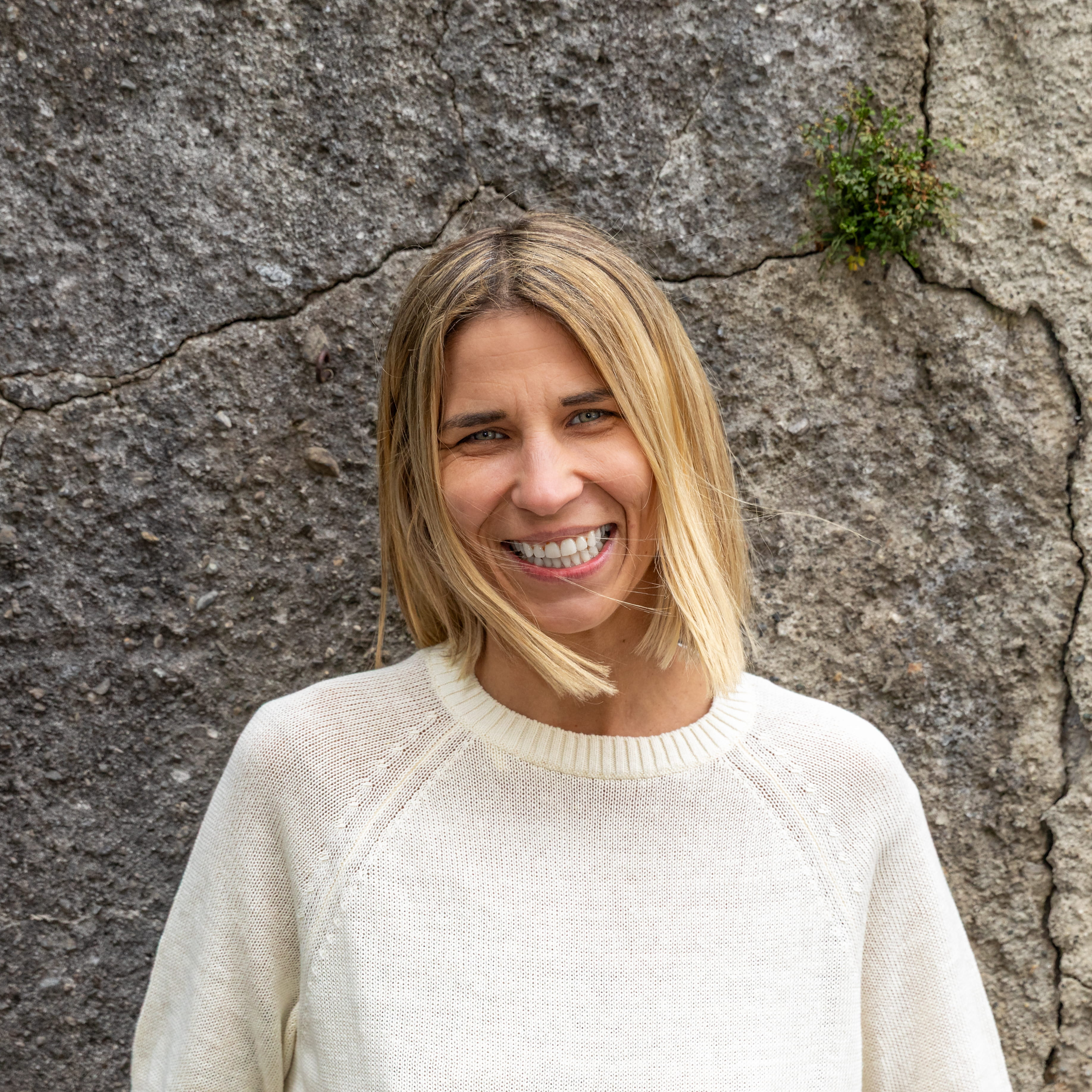 Janine steht lächelnd vor einer Steinmauer. Sie hat blonde, kinnlange Haare und ein weißes Oberteil.